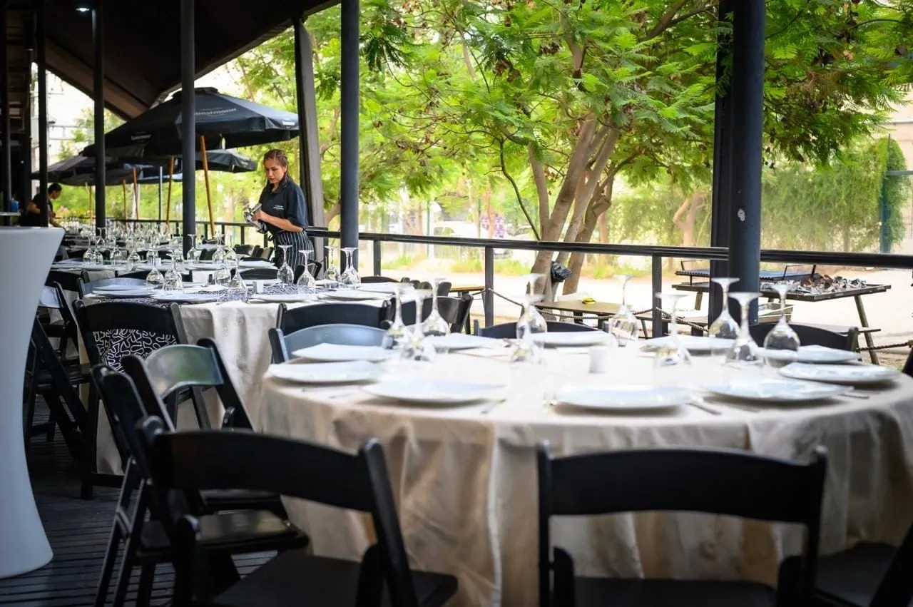 La terraza de La Miga montada para un evento, con mesas redondas vestidas, copas y flores bajo los árboles