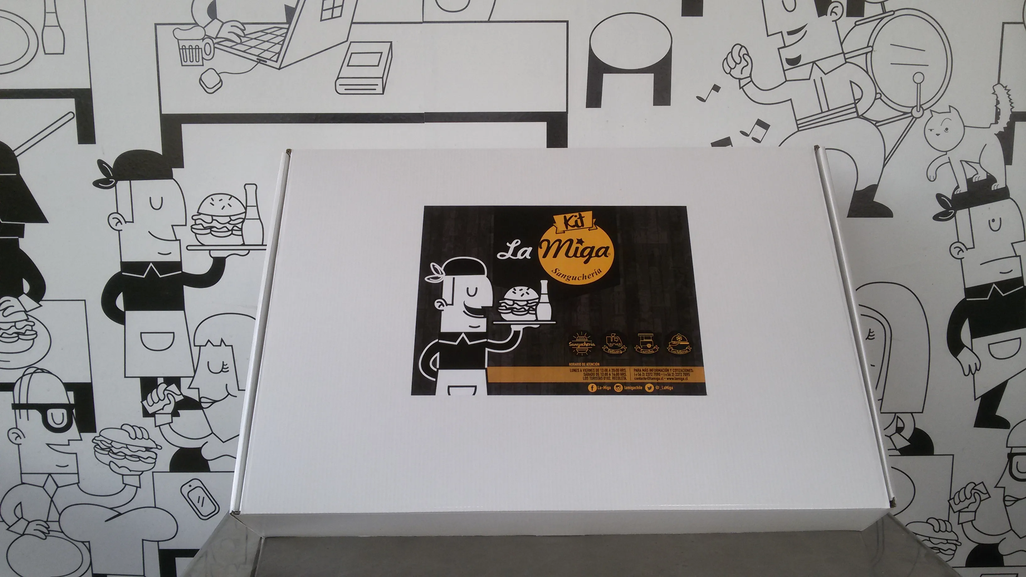 Caja del Kit La Miga sobre una mesa, con el mural de personajes del local de fondo