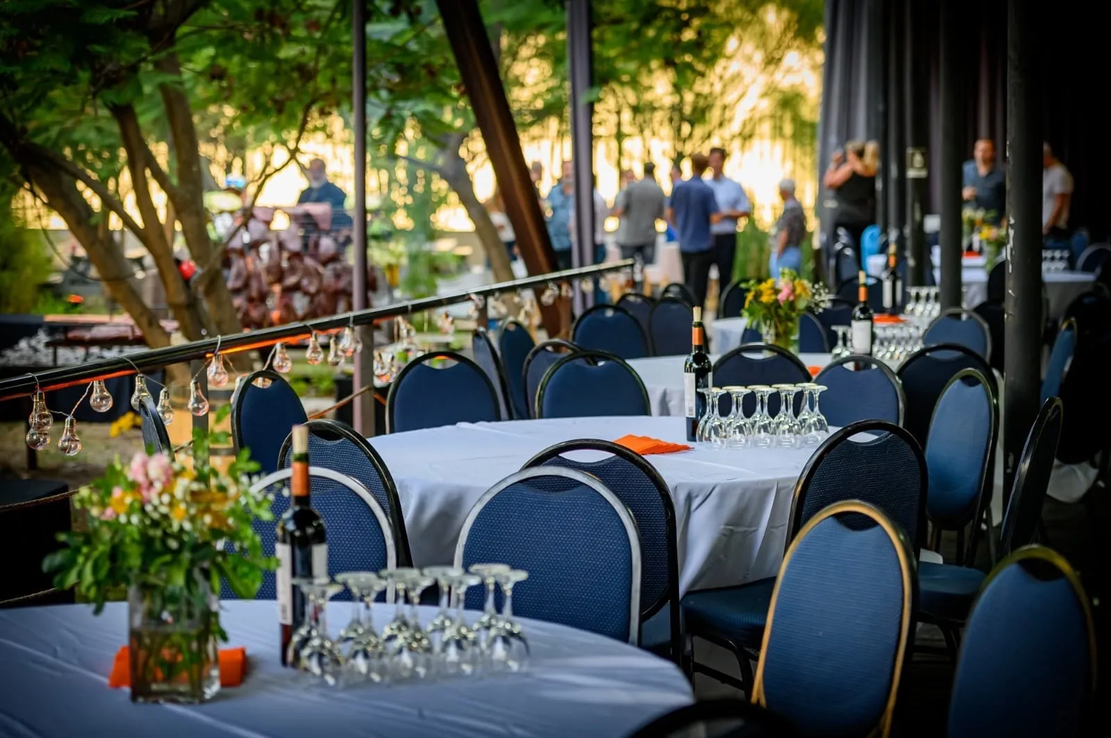 Mesas redondas montadas para un banquete, con mantel, copas y sillas azules, bajo los árboles de la terraza